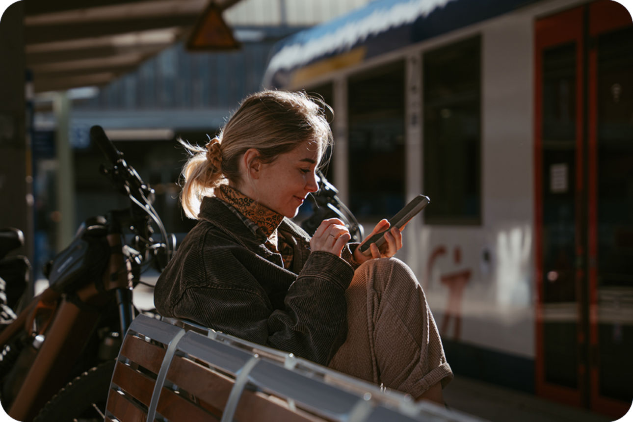 Person sitzt mit Handy am Bahnsteig und wartet auf den Zug.