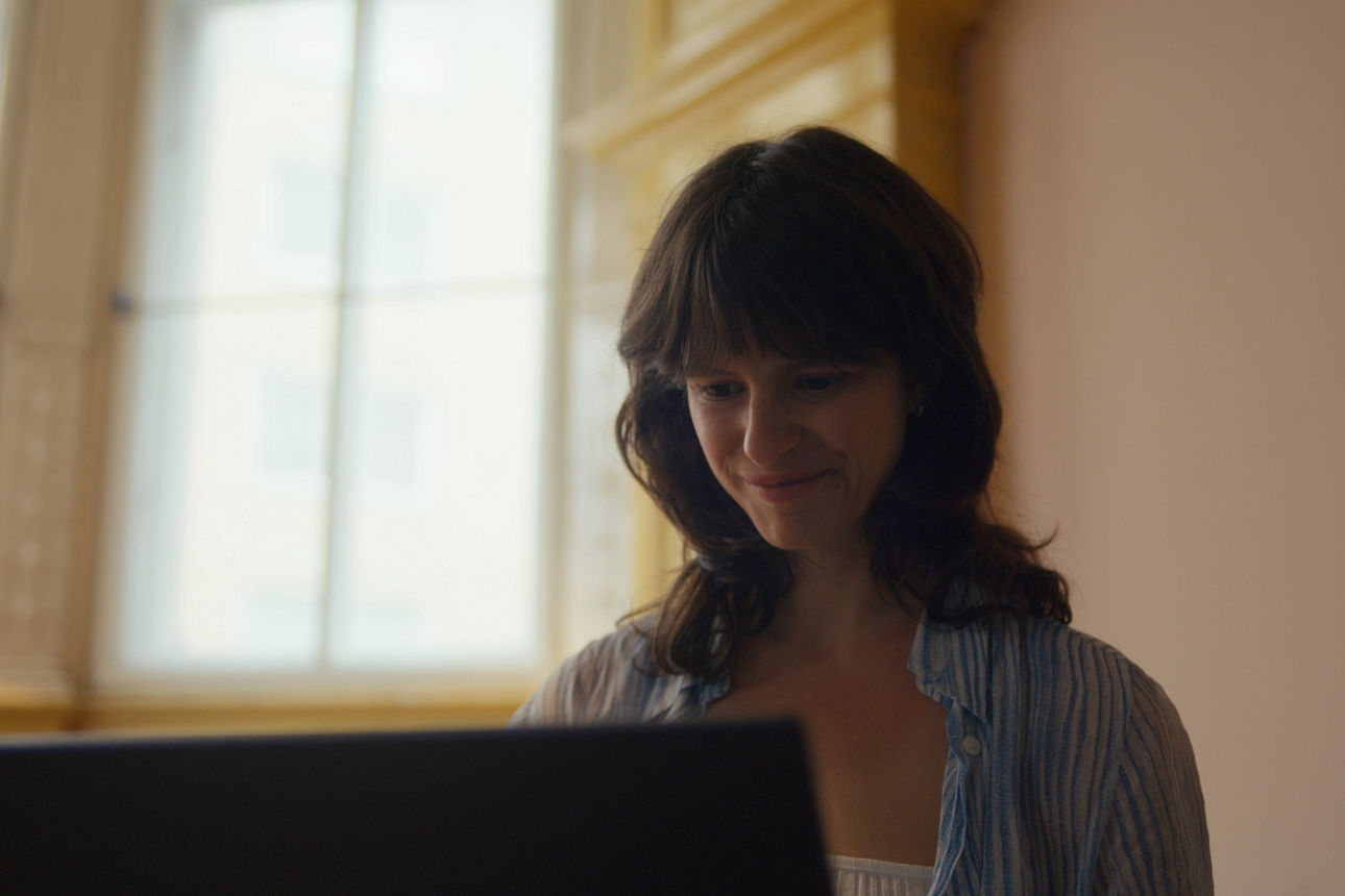 Im Vordergrund sitzt eine Frau am Laptop. Im Hintergrund erkennt man eine Wohnung mit heller Fensterfront. Im Vordergrund sitzt eine Frau am Laptop. Im Hintergrund erkennt man eine Wohnung mit heller Fensterfront.
