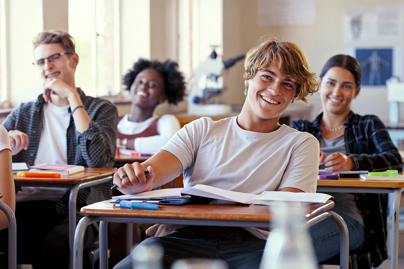 Schülerinnen und Schüler sitzen in einem Klassenraum und lächeln in die Kamera