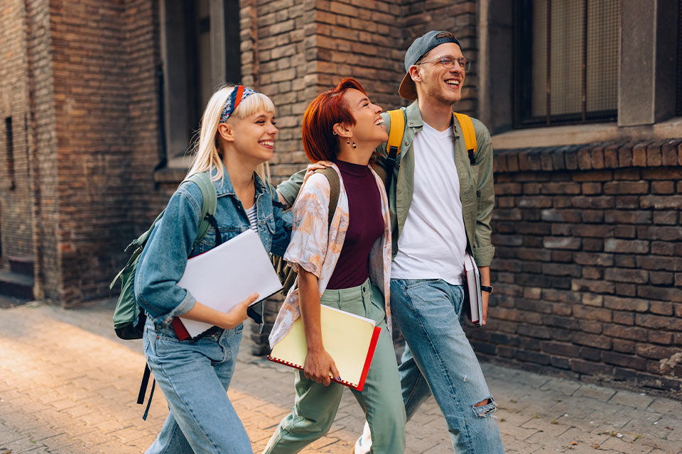 Zwei Schülerinnen und ein Schüler laufen lachend vor einem Gebäude entlang