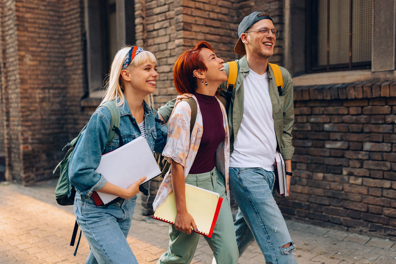 Zwei Schülerinnen und ein Schüler laufen lachend vor einem Gebäude entlang