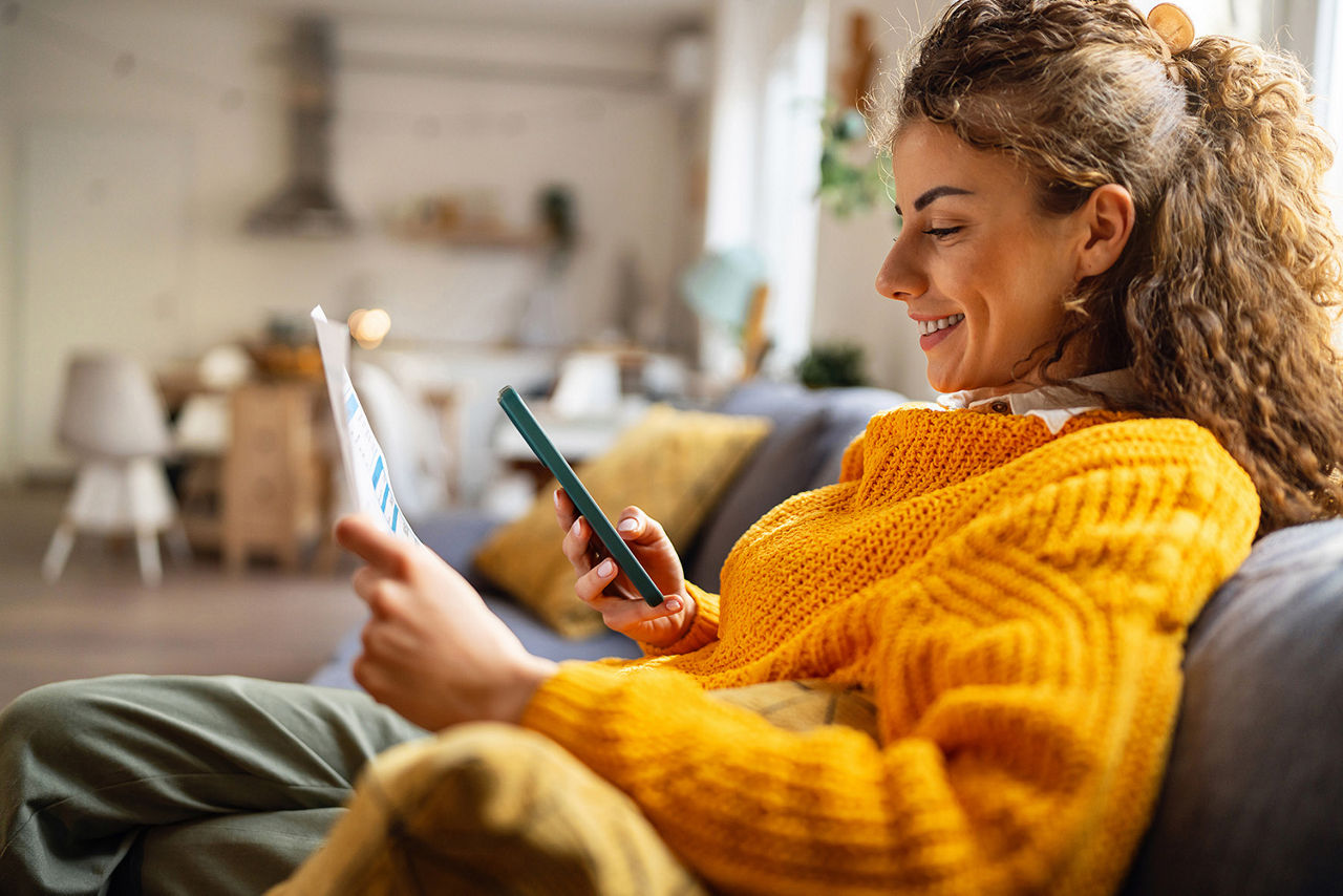 Junge Frau sitzt auf der Couch mit Ihrem Handy und hat einen Brief in der Hand