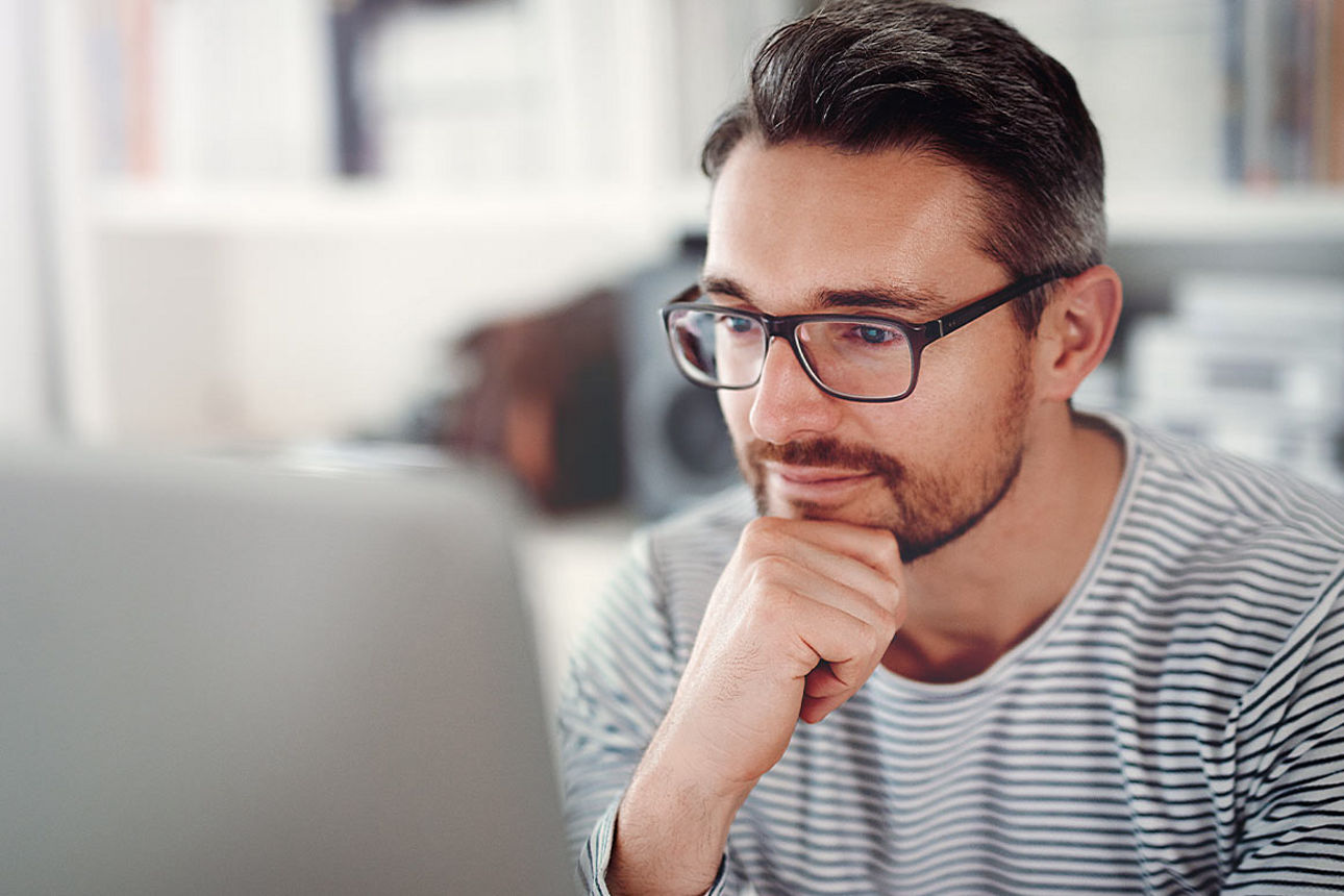 Mann mit Brille sitz vor einen Laptop und hält mit der Hand sein Kinn
