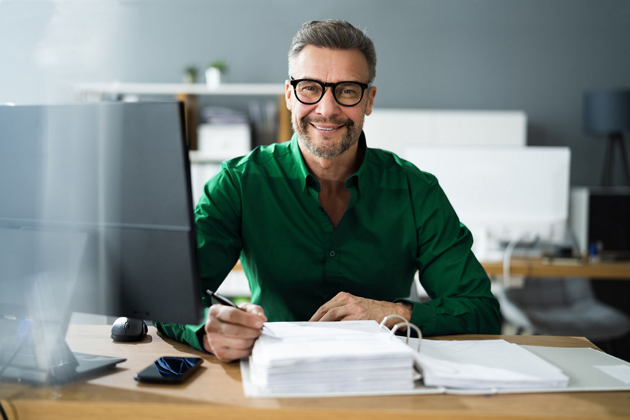 Ein Mann mit grünem Hemd und Brille sitzt an einem Schreibtisch.