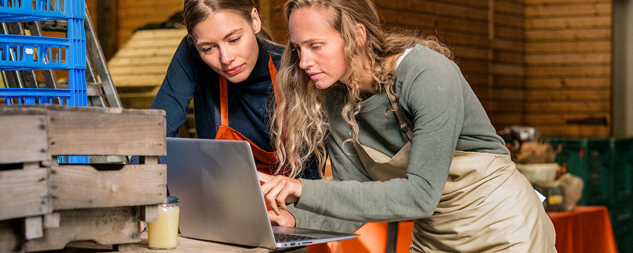 Zwei junge Frauen schauen gemeinsam auf einen Laptop und stehen in einem Veranstaltungsraum