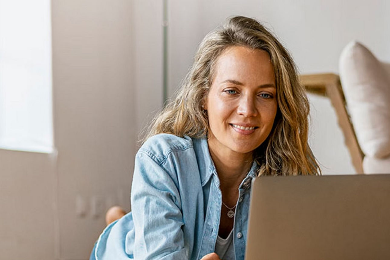 Junge Frau liegt gemütlich auf dem Boden ihres Wohnzimmers und schaut lächelnd in ihr Laptop 