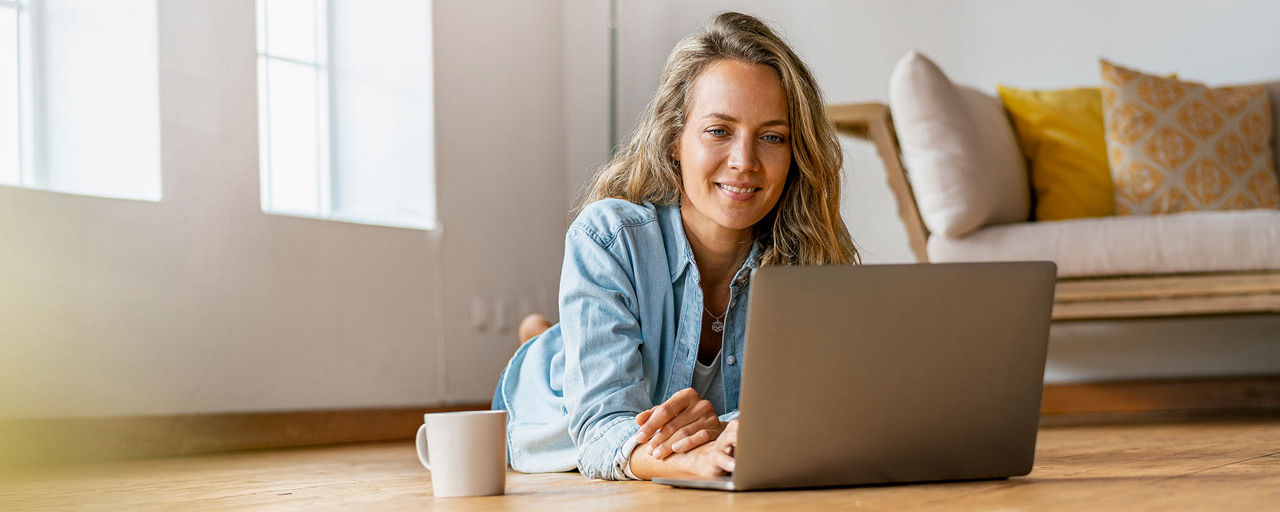 Eine junge Frau liegt im Wohnzimmer auf dem Boden und guckt lächelnd auf ihren Laptop