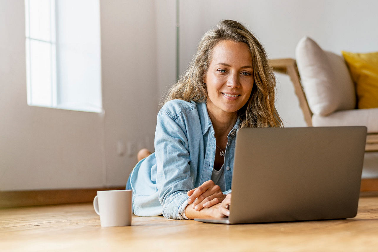 Eine Frau liegt entspannt auf dem Boden ihres Wohnzimmers und arbeitet am Laptop