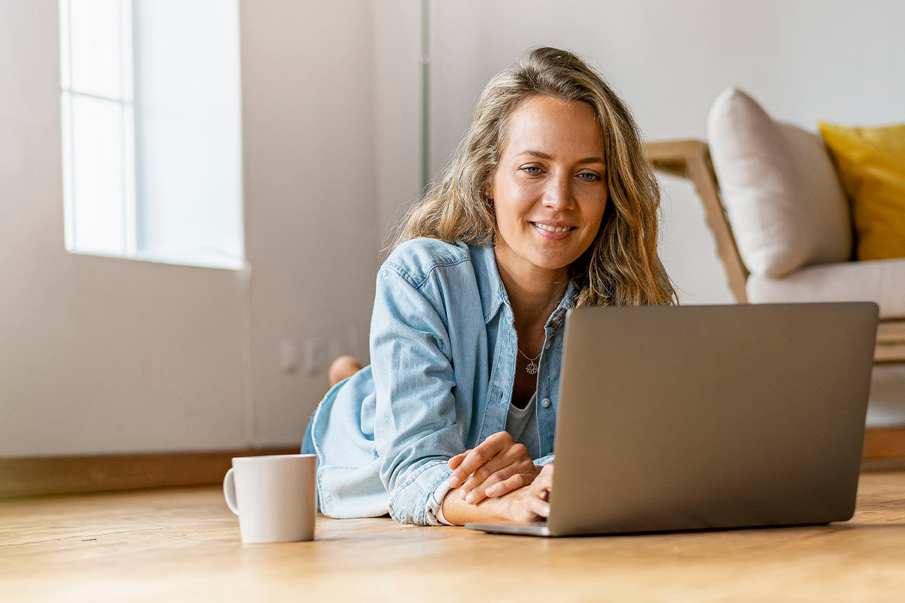 Eine Frau liegt entspannt auf dem Boden ihres Wohnzimmers und arbeitet am Laptop