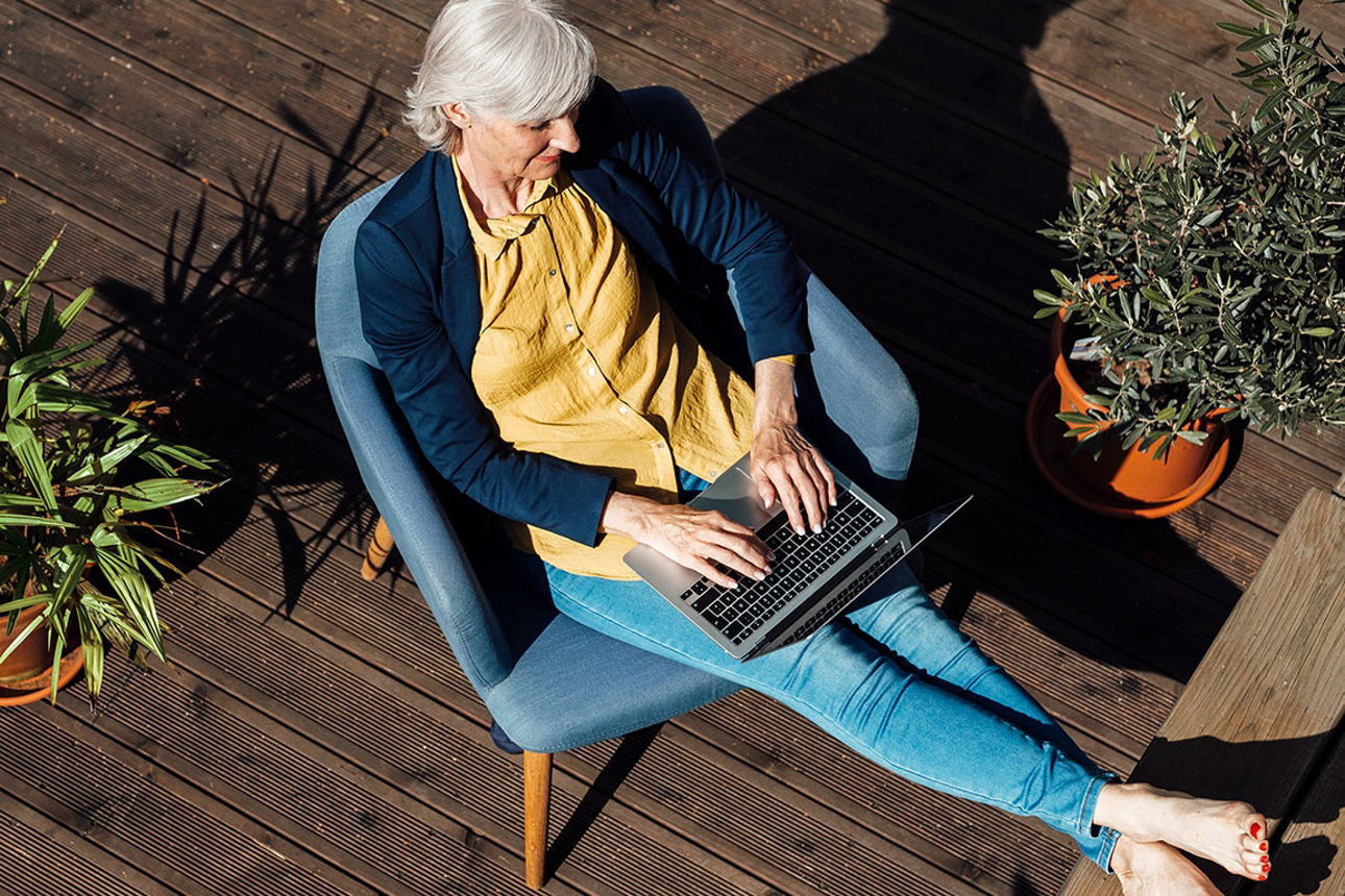 Eine Frau sitzt entspannt auf ihrer Terrasse und hat einen Laptop auf dem Schoß.