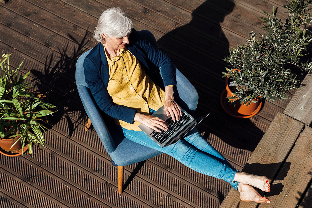 Eine ältere Frau sitzt entspannt auf einer Terrasse und arbeitet am Laptop