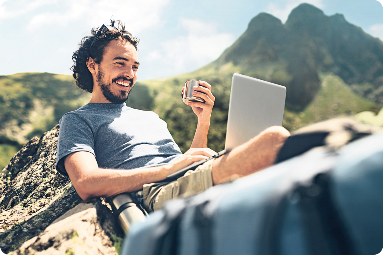 Ein Mann sitzt lächelnd auf einem Felsen mit einem Laptop auf dem Schoß, hält eine Tasse in der Hand und im Hintergrund sind Berggipfel zu sehen