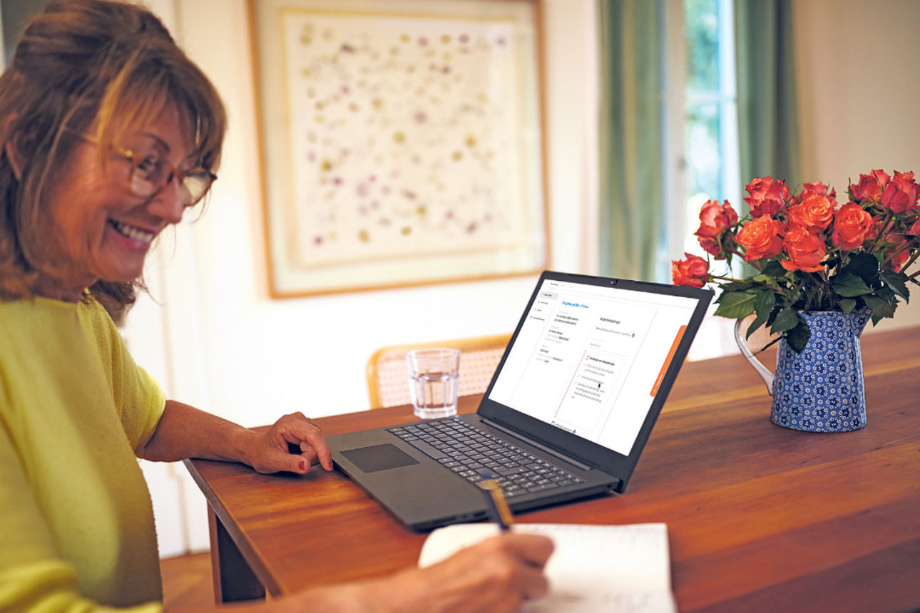 Eine Frau sitzt mit einem Laptop am Küchentisch und macht sich Notizen beim OnlineBanking.