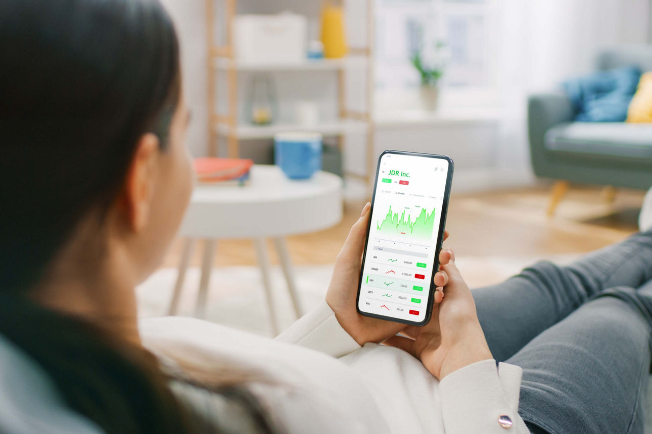 Junge Frau sitzt zu Hause auf der Couch und nutzt Smartphone mit Börsen-App, um Aktienkurse zu prüfen. Die Kurve steigt und erzielt einen hohen Gewinn. Im Hintergrund gemütliche Atmosphäre.