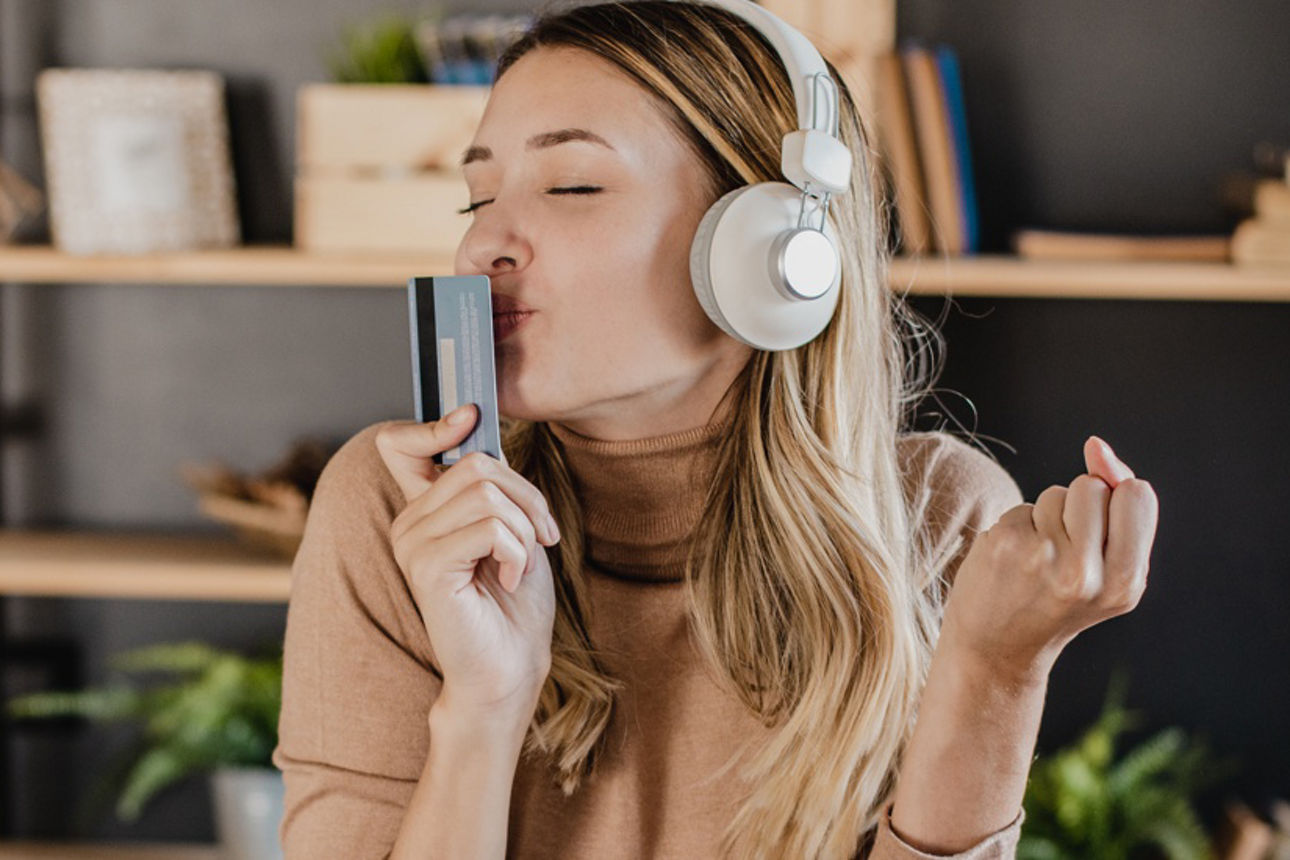 Junge Frau sitzt mit Kopfhörern vor einem Notebook und küsst ihre Kreditkarte