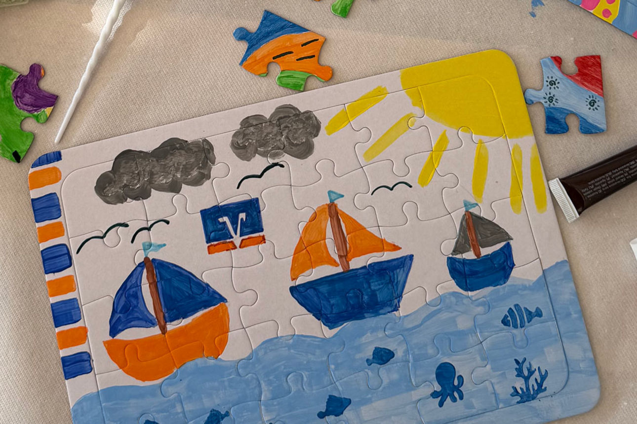 Ein bemaltes Puzzle mit Segelschiffen auf dem Meer, umgeben von Farbtuben, Malkasten und Puzzleteilen.