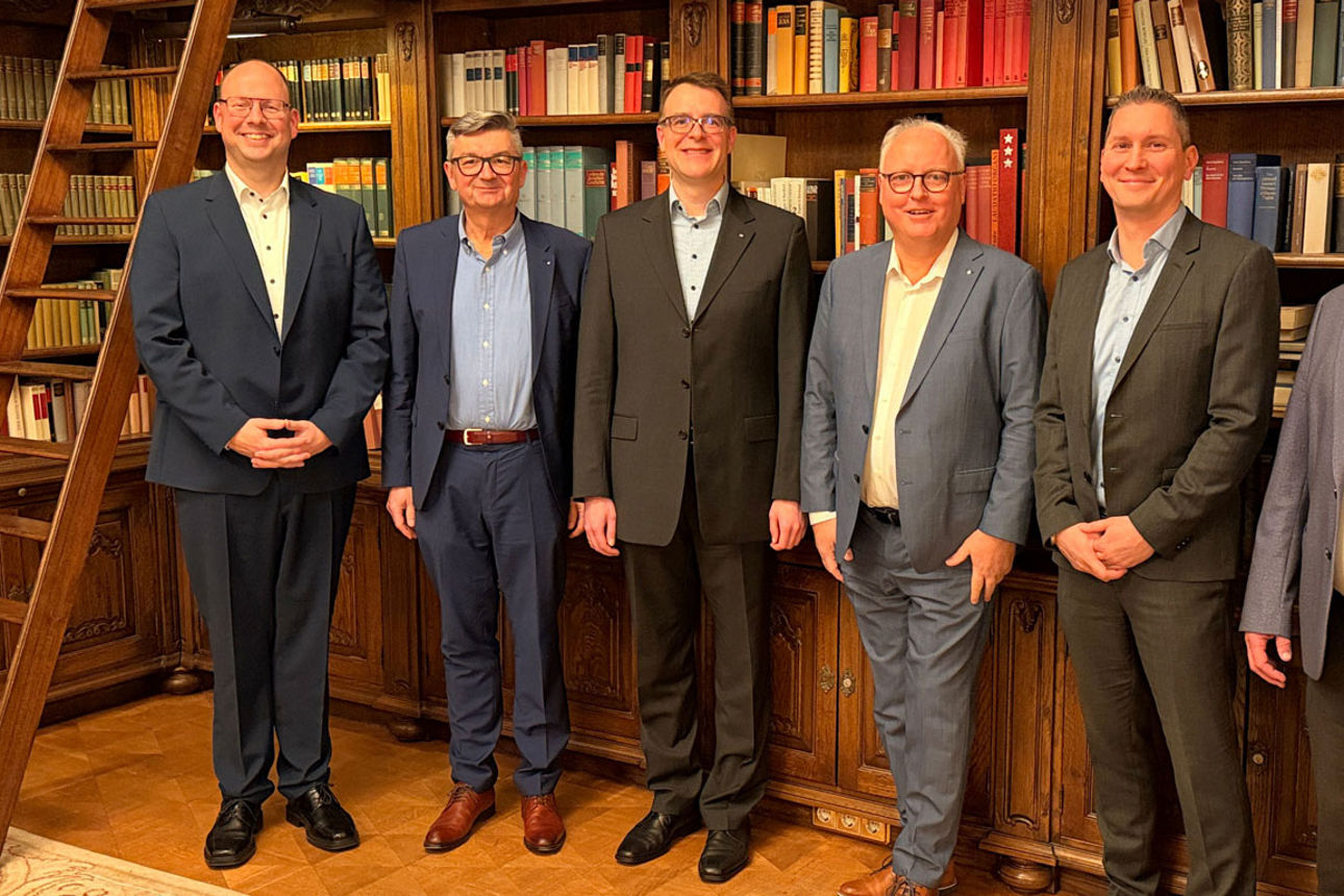 Sechs Männer stehen in einer Bibliothek vor gefüllten Bücherregalen und posieren gemeinsam für ein Gruppenfoto.