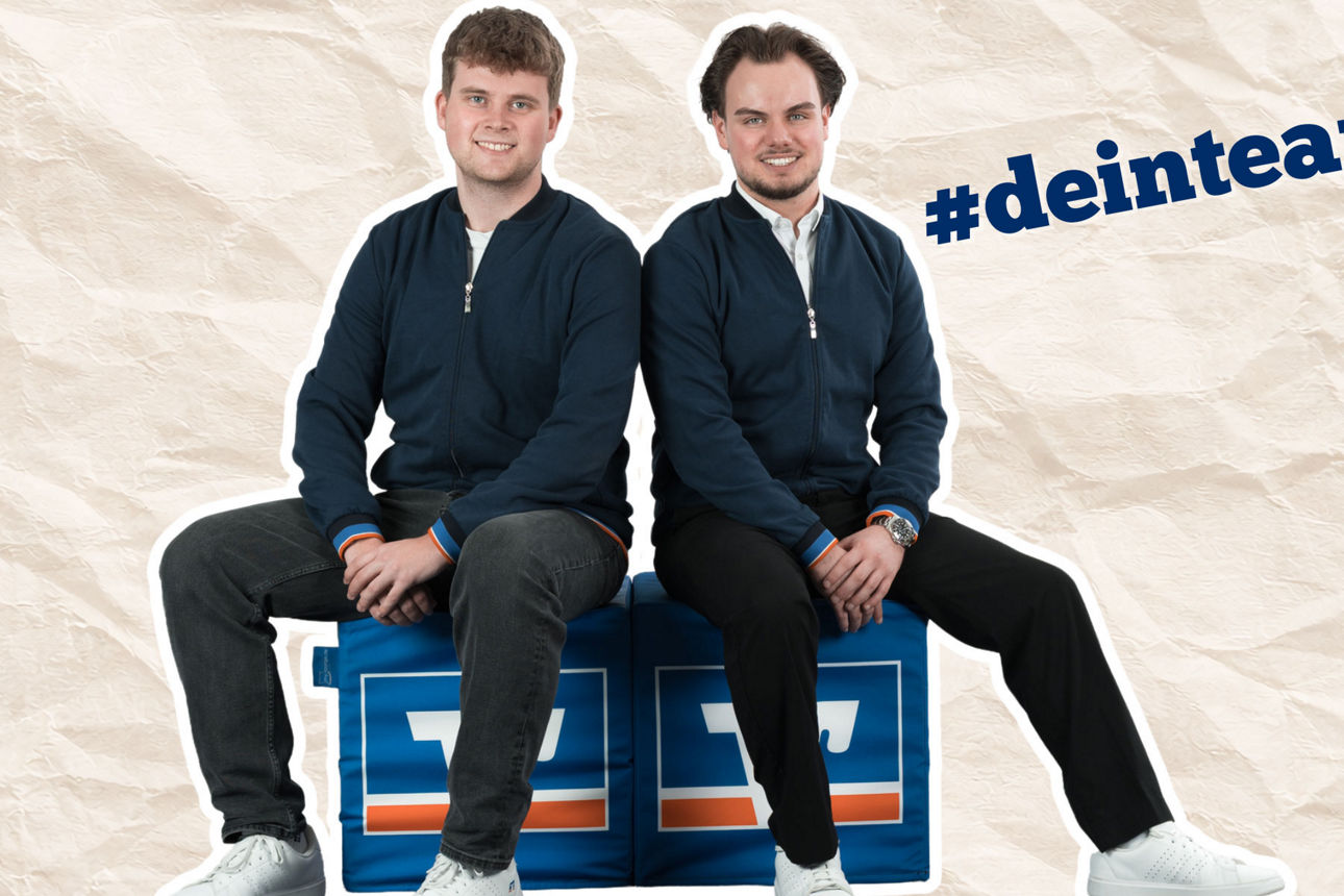 Zwei Männer sitzen Rücken an Rücken auf blauen VR-Bank-Sitzwürfeln vor hellem Hintergrund.