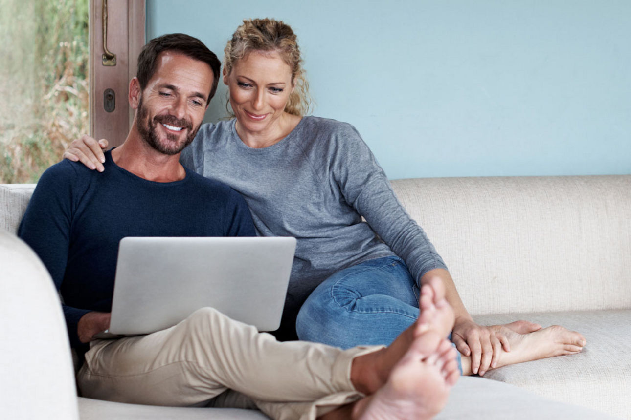 Ein entspanntes Paar sitzt gemeinsam auf einem hellen Sofa in einem modernen Wohnzimmer und blickt lächelnd auf einen Laptop.