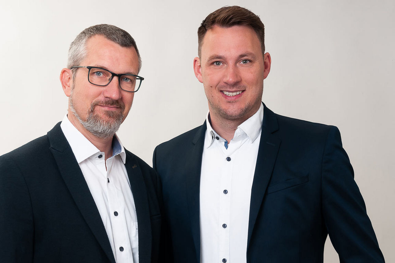Zwei sympathische Geschäftspartner präsentieren sich gemeinsam vor einem hellen Hintergrund.