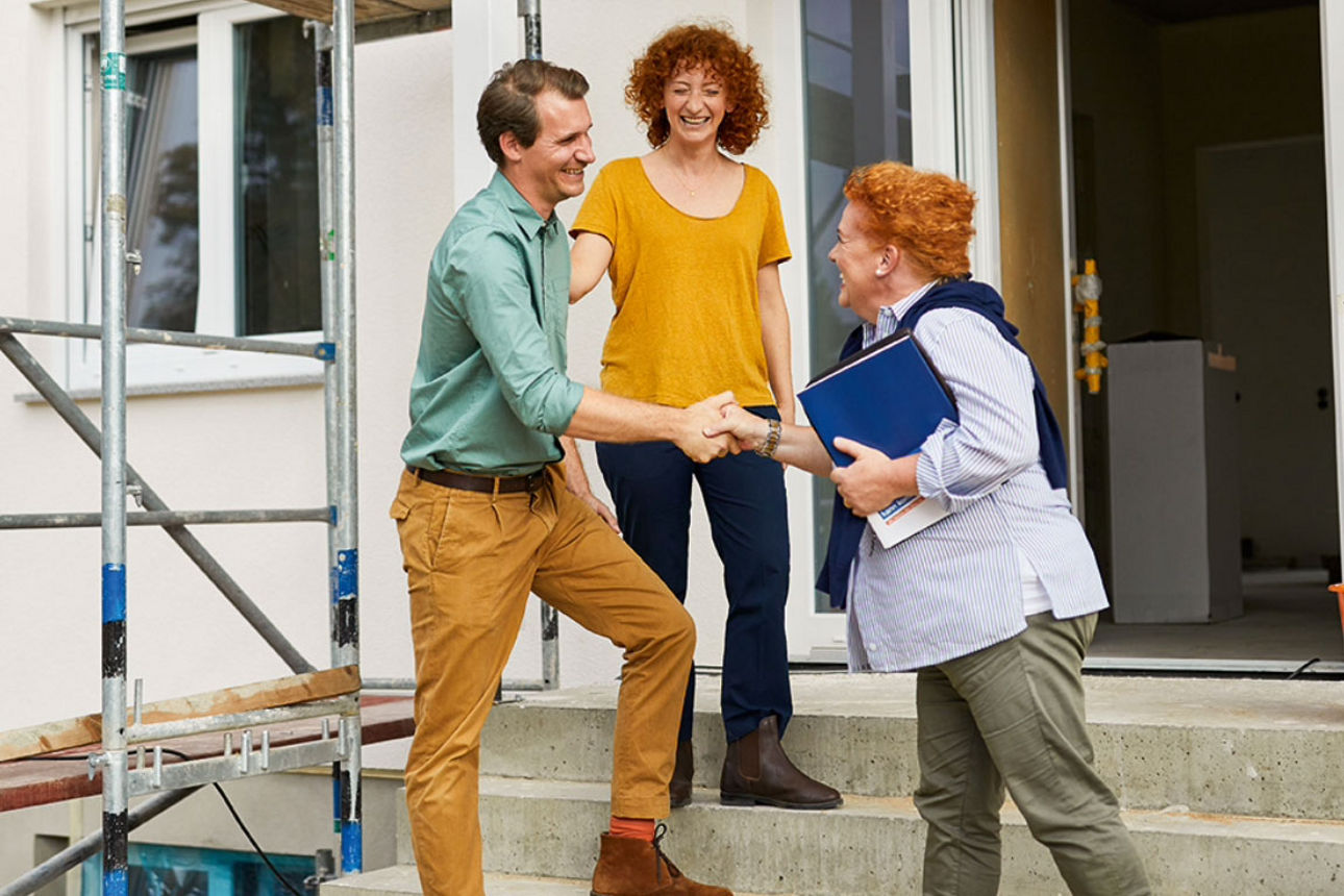 Gemini hat gesagt  Ein Paar und eine Beraterin geben sich vor einem Haus im Bau auf einer Baustelle mit Gerüst die Hände.