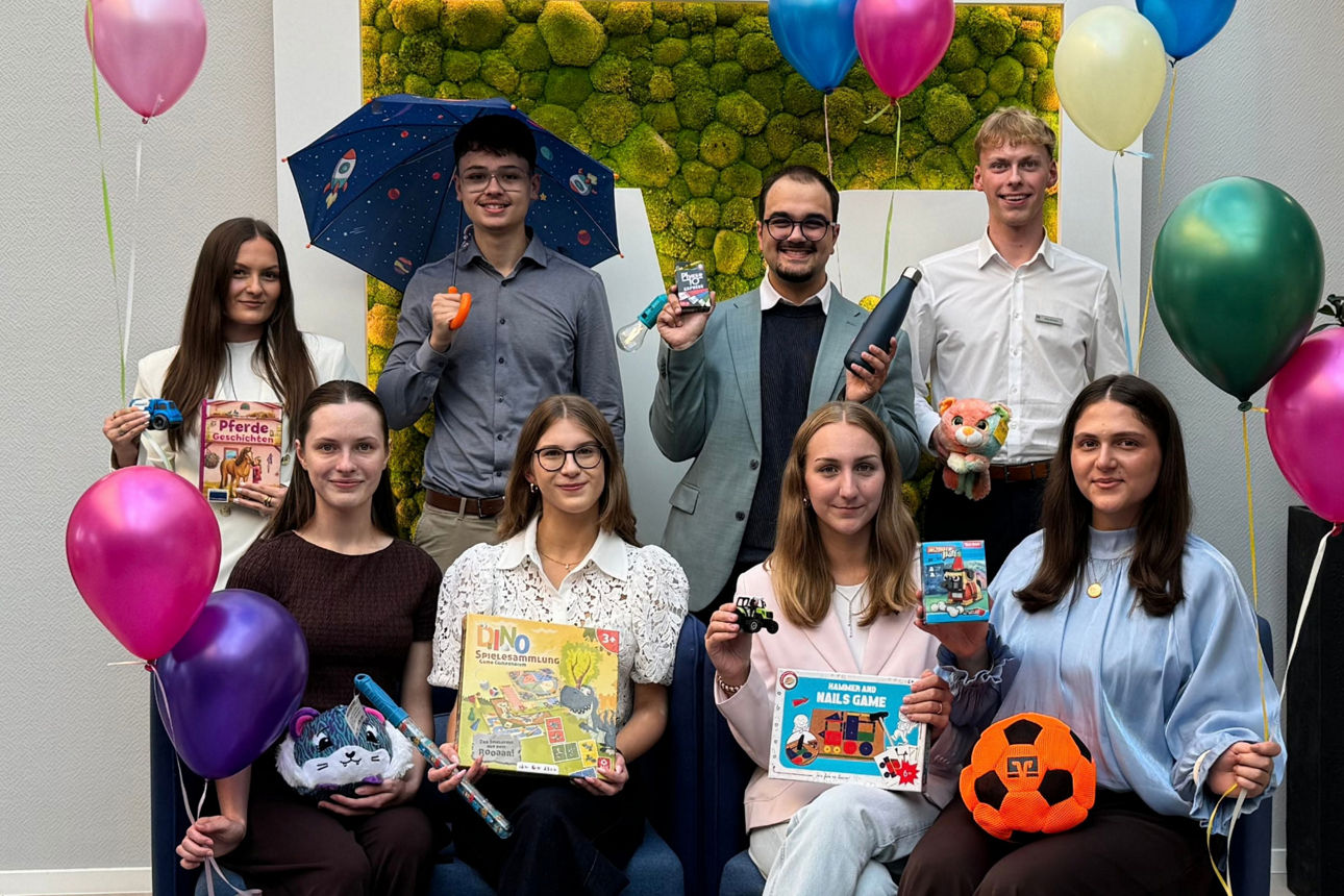 Eine fröhliche Gruppe von Personen posiert mit Geschenken und bunten Luftballons vor einer dekorativen Pflanzenwand.