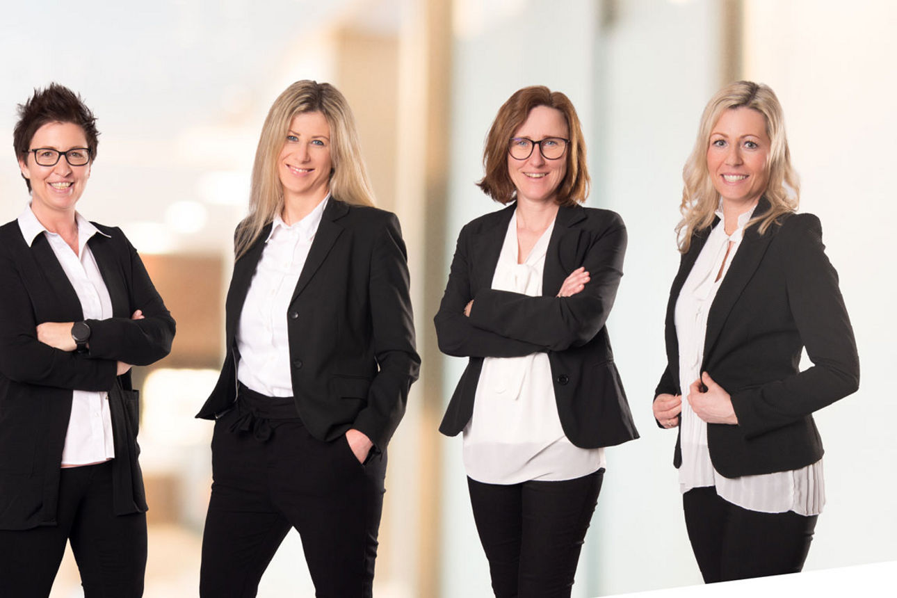 Sechs lächelnde Frauen in professioneller Business-Kleidung (schwarze Blazer, weiße Oberteile) posieren selbstbewusst in eine