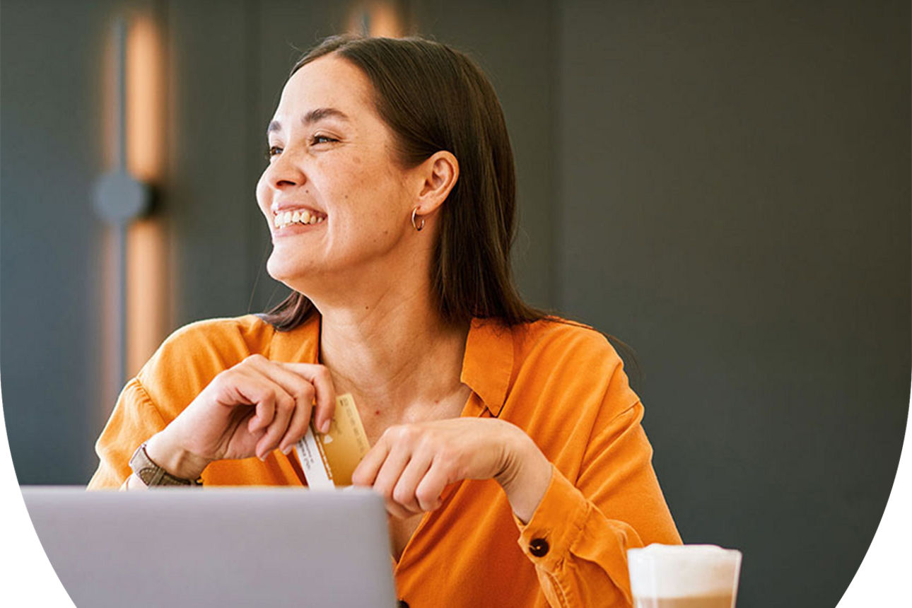 Eine Frau sitzt mit Laptop und Kaffee an einem Tisch und hält ihre goldene girocard in den Händen