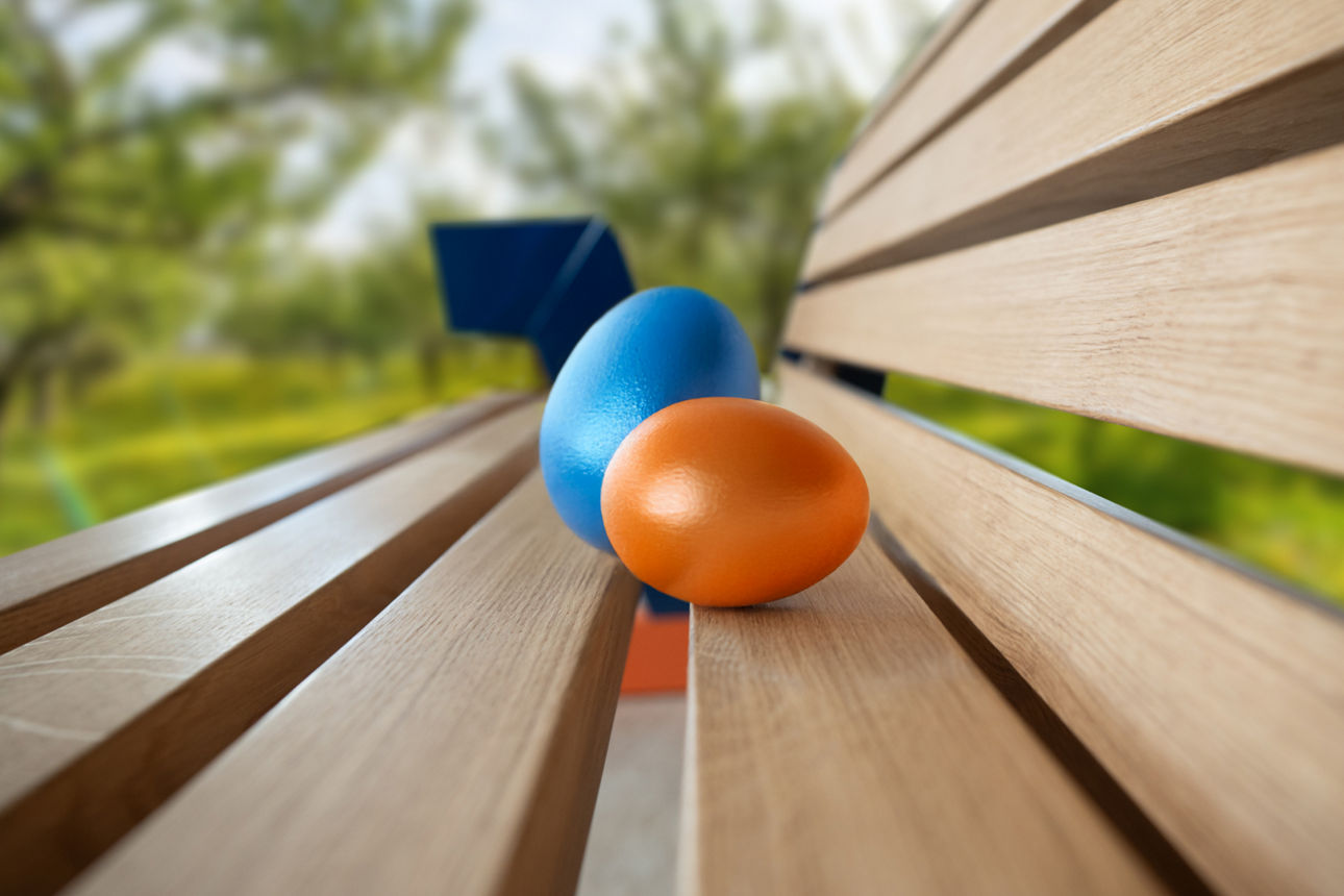 Zwei Ostereier in Blau und Orange auf einer Holzbank im Grünen.