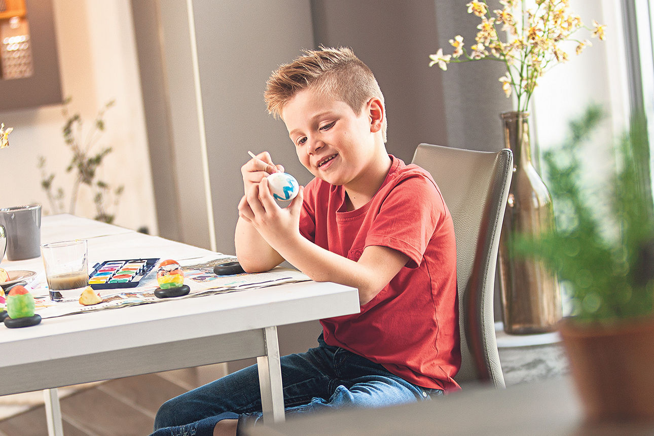 Junge mit rotem T-Shirt sitzt am Tisch und malt Eier an