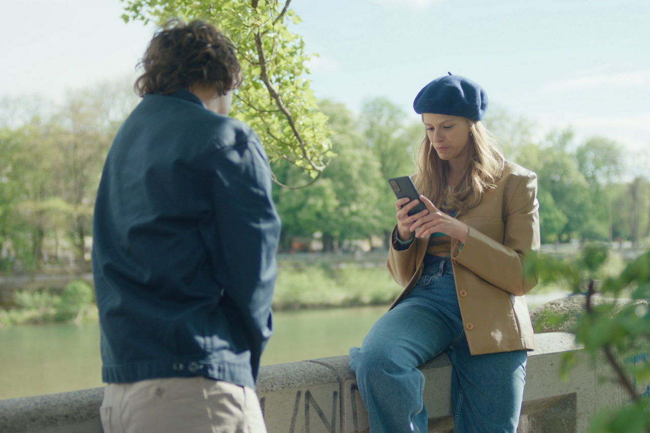 Ein junges Paar sitzt auf einer Steinmauer an einem See im Park. Die Frau schaut gerade auf ihr Handy, der Mann schaut auf den See.