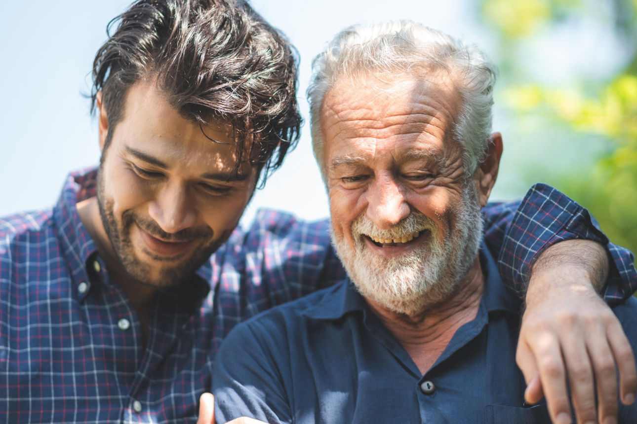 Zwei Personen sitzen eng beieinander im Grünen und legen fürsorglich die Arme umeinander.