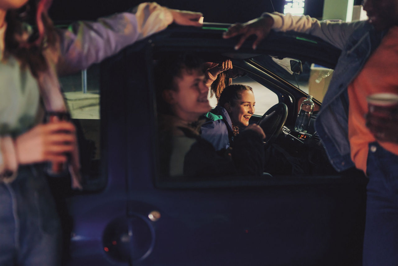 Endlich 18 - Zwei Jugendliche sitzen lachend im parkendem Auto und drei Freunde stehen daneben
