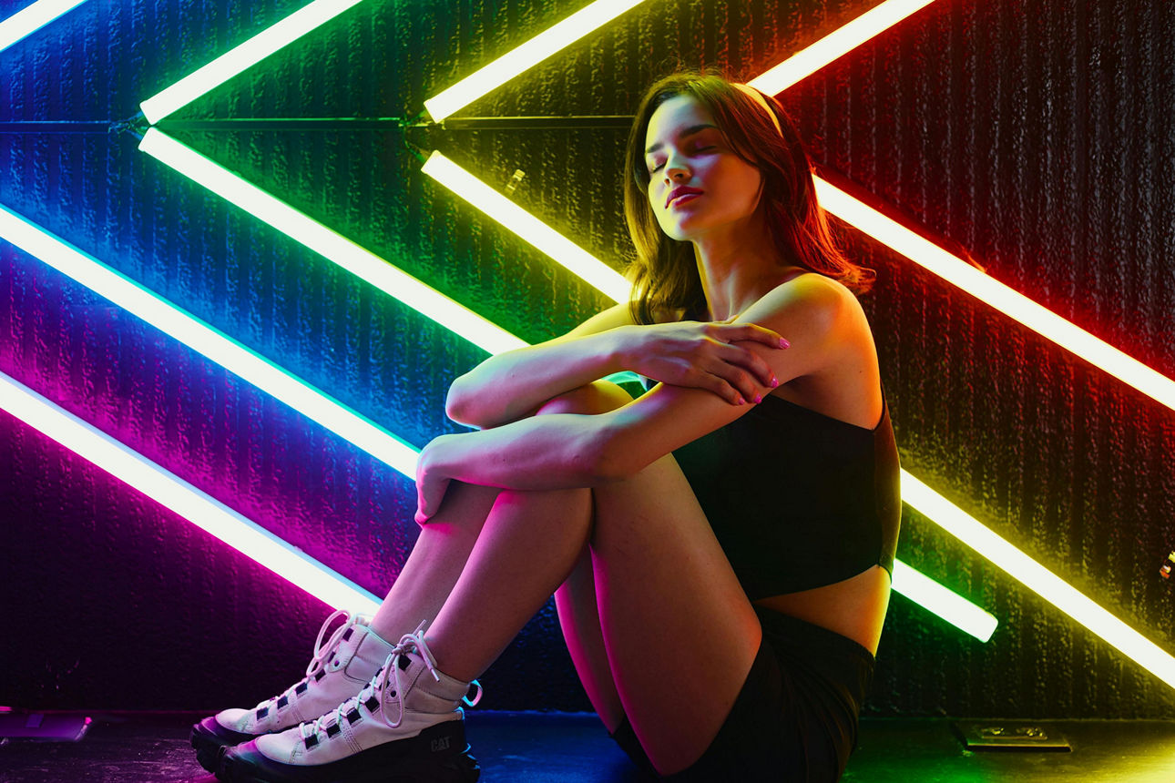 Junge Frau sitzt vor diagonalen Neonröhren in bunten Farben und wird von farbigem Licht beleuchtet.