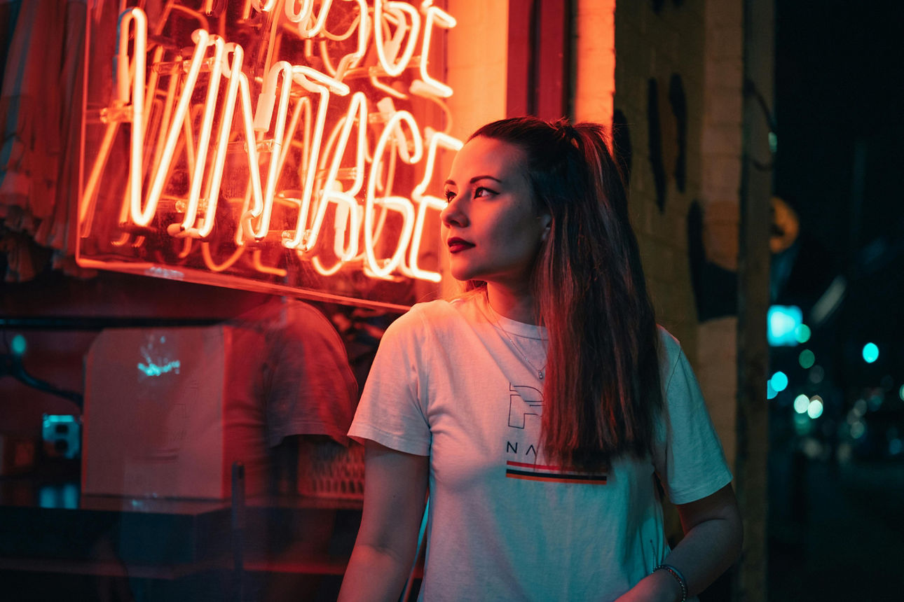 Frau mit weißem T-Shirt steht bei Nacht vor einem orangefarbenen Neon-Vintage-Schild.
