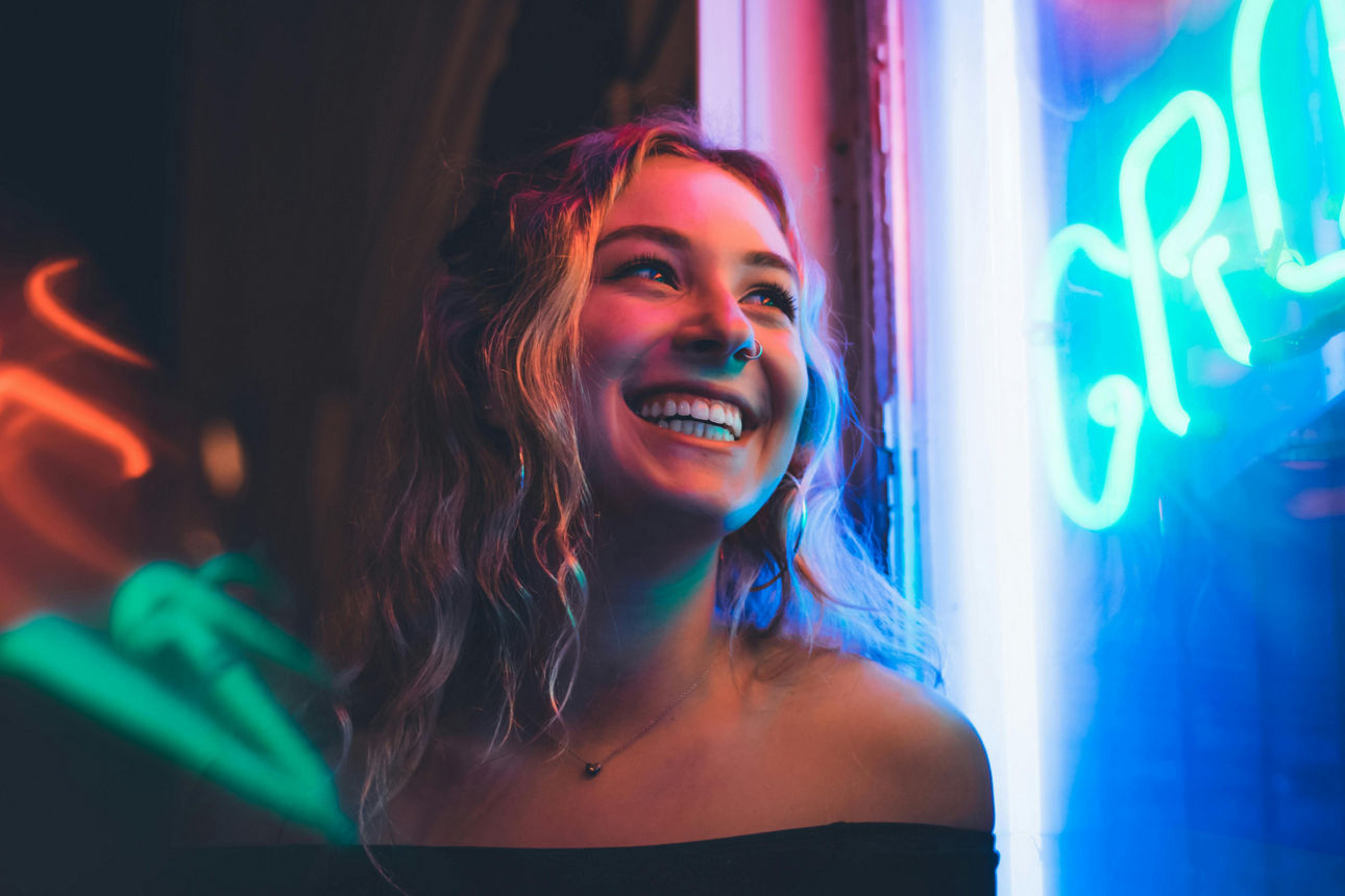 Lächelnde Frau mit offenen Haaren steht vor einem Fenster mit blauem und pinkem Neonlicht.