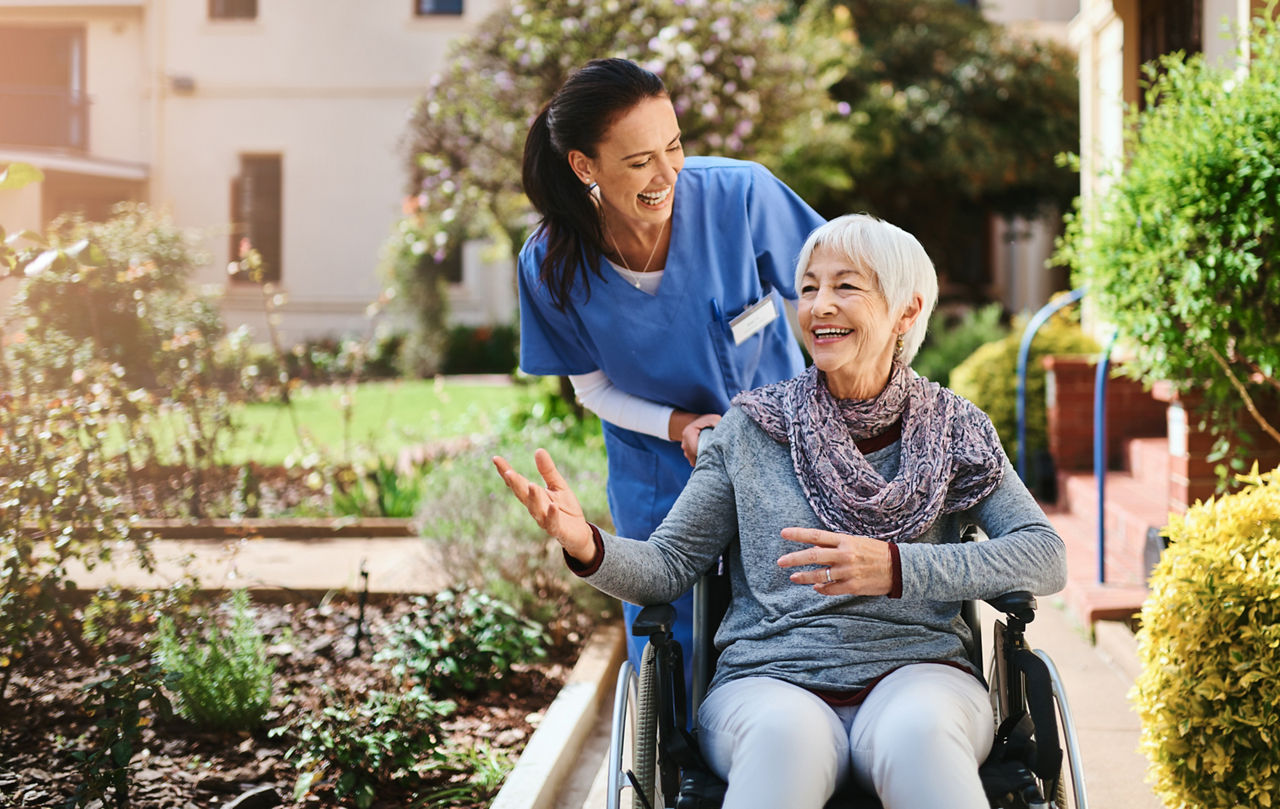 Krankenschwester schiebt Seniorin im Rollstuhl im Park, beide lachen und reden miteinander