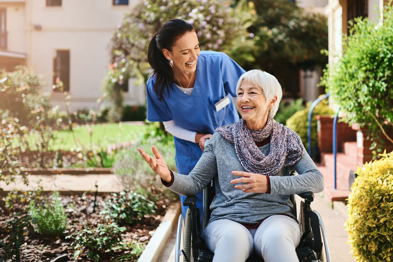 Krankenschwester schiebt Seniorin im Rollstuhl im Park, beide lachen und reden miteinander