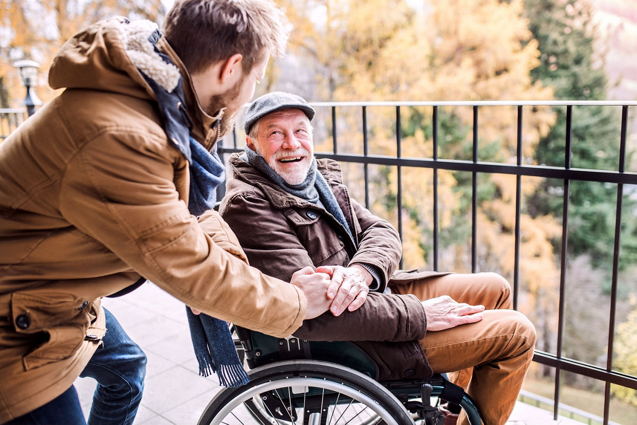 Ein junger Mann steht seitlich, leicht nach vornge gebeugt neben einem älteren Herren im Rollstuhl. Sie lächeln sich an. Der Ältere hat dem Jüngeren seine Hand auf dessen Hand gelegt. 