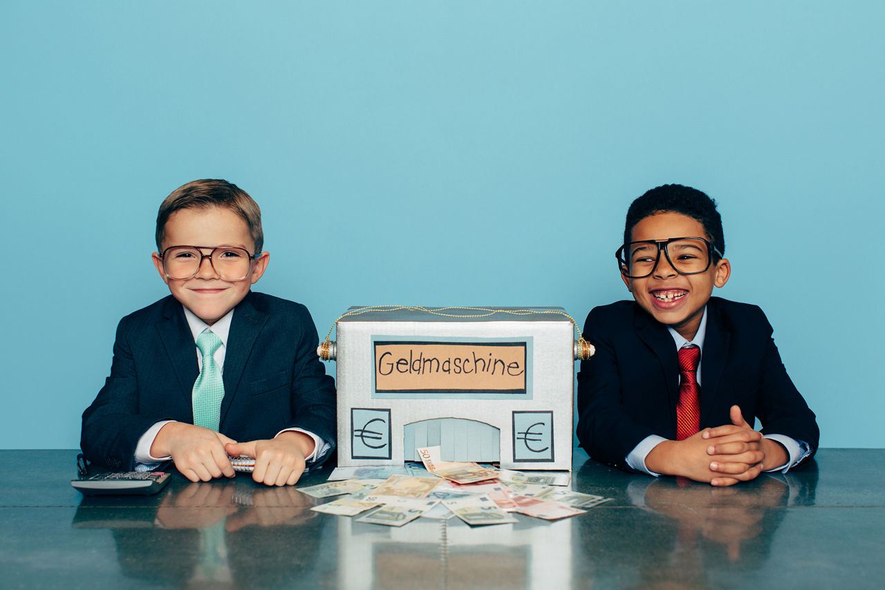 Zwei Jungen mit Anzug und Krawatte, in der Mitte steht eine Box mit der Aufschrift Geldmaschine.
