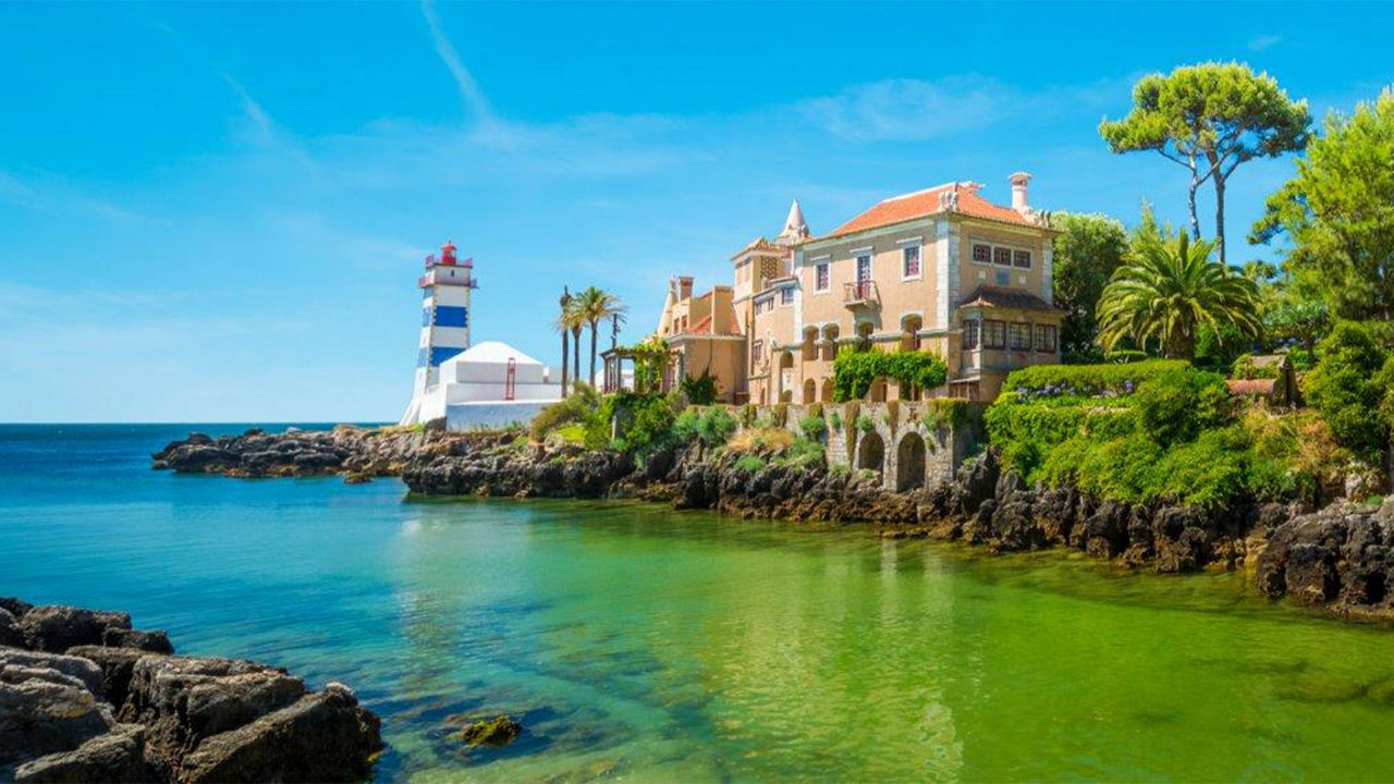 Bucht von Cascais mit türkis-blauem Meer und einer Landzunge mit Leuchtturm und mediterrandem Gebäude