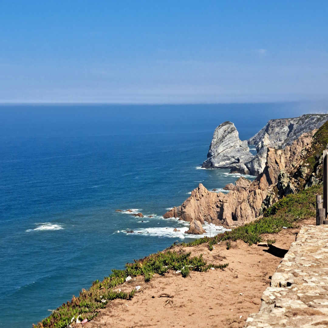 Blick über eine Klippe auf offenes Meer und schroffe verschiedenfarbige Felsen, an denen sich die Wellen brechen