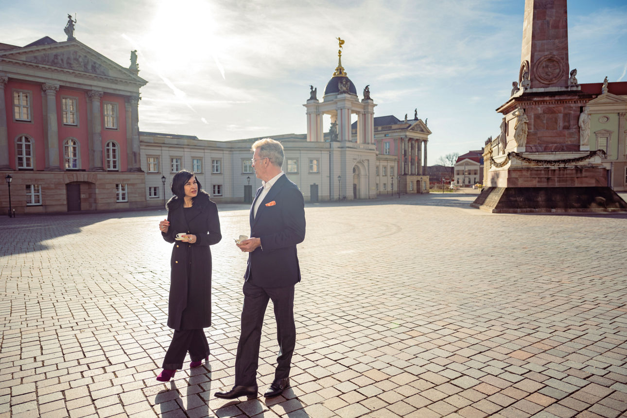 Potsdam in Wandel Zwei Geschäftsleute gehen mit Kaffee über einen historischen Platz und unterhalten sich.