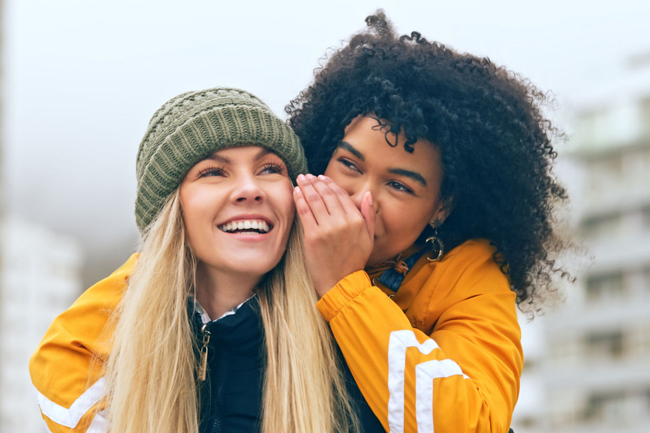 Zwei junge Frauen - eine flüstert der anderen was ins Ohr
