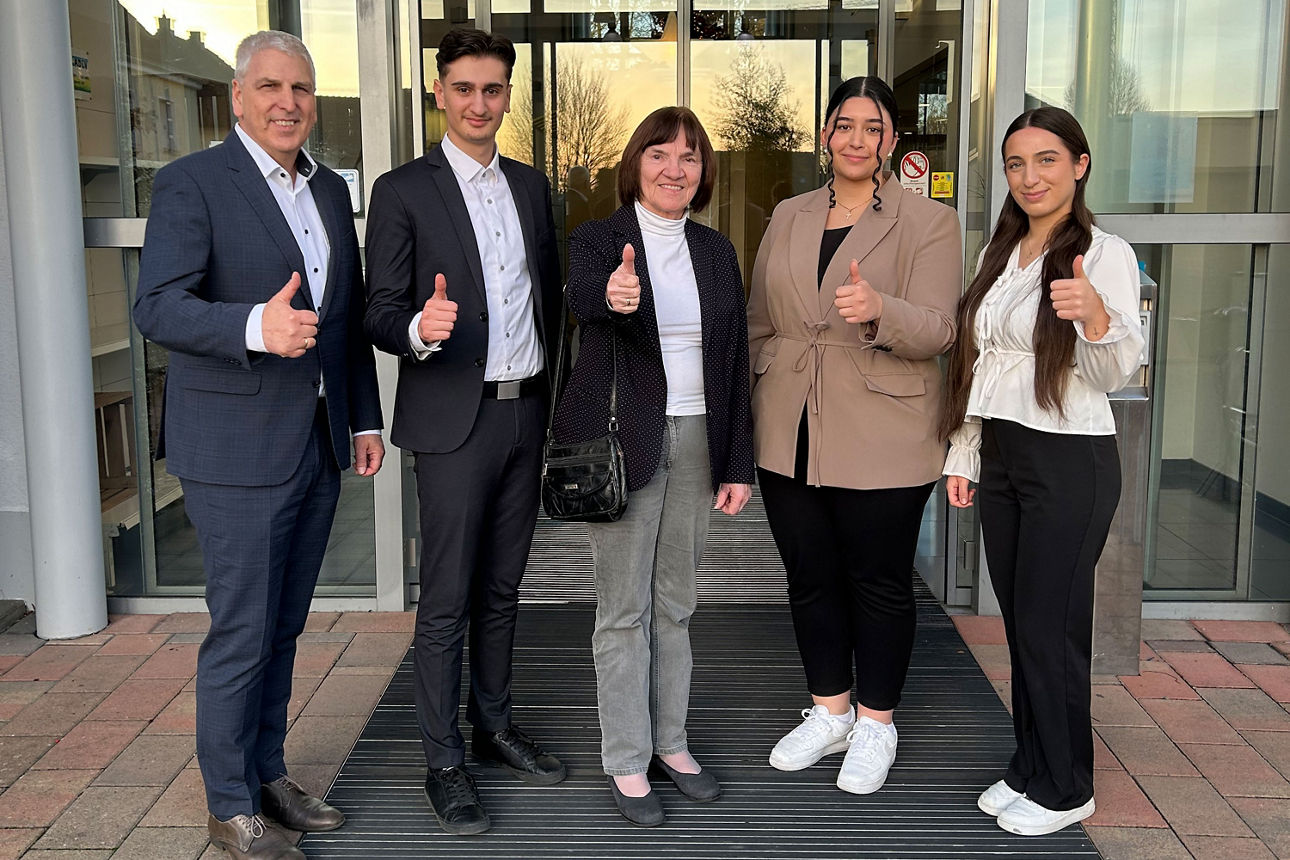 Fünf Personen stehen vor einem Gebäude, lächeln in die Kamera und zeigen geschlossen den Daumen nach oben.