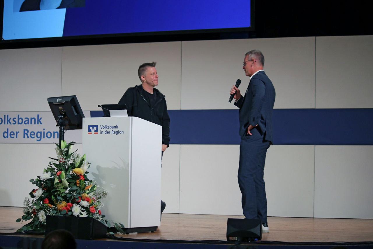 Vorstand Thomas Taubenberger hält ein Mikrofon und spricht auf der Bühne mit dem Referenten Linus Neumann.
