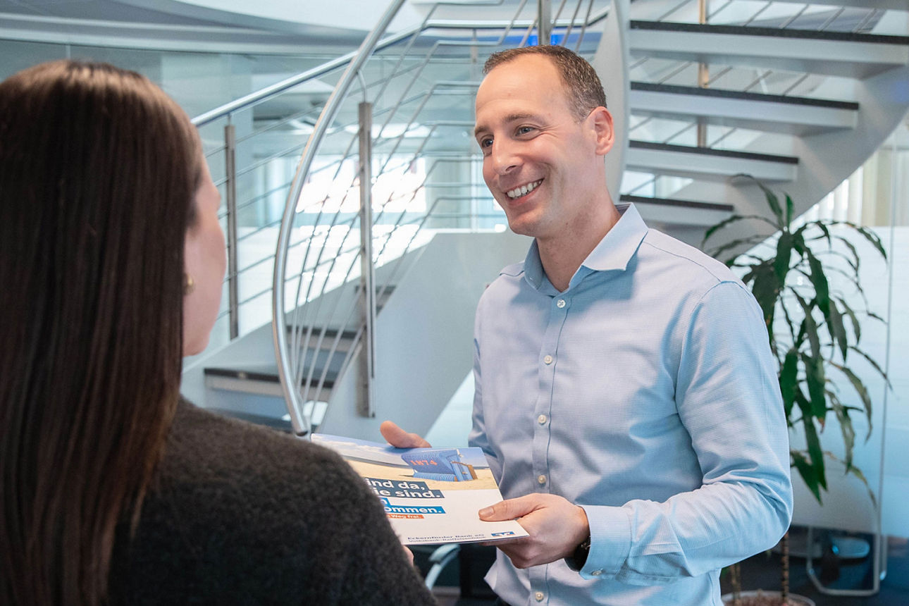 Bankberater überreicht Unterlagen im Gespräch mit Kundin, freundliche Beratungssituation im modernen Büro.