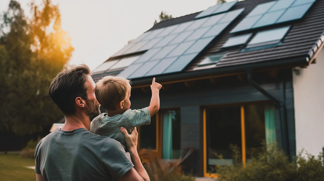 Vater steht mit seinem Sohn auf dem Arm vor einem Haus mit Solarpanelen auf dem Dach.