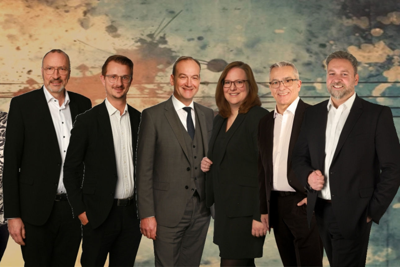 Gruppe elegant gekleideter Personen posiert vor einem farbigen Hintergrund in Business-Atmosphäre.