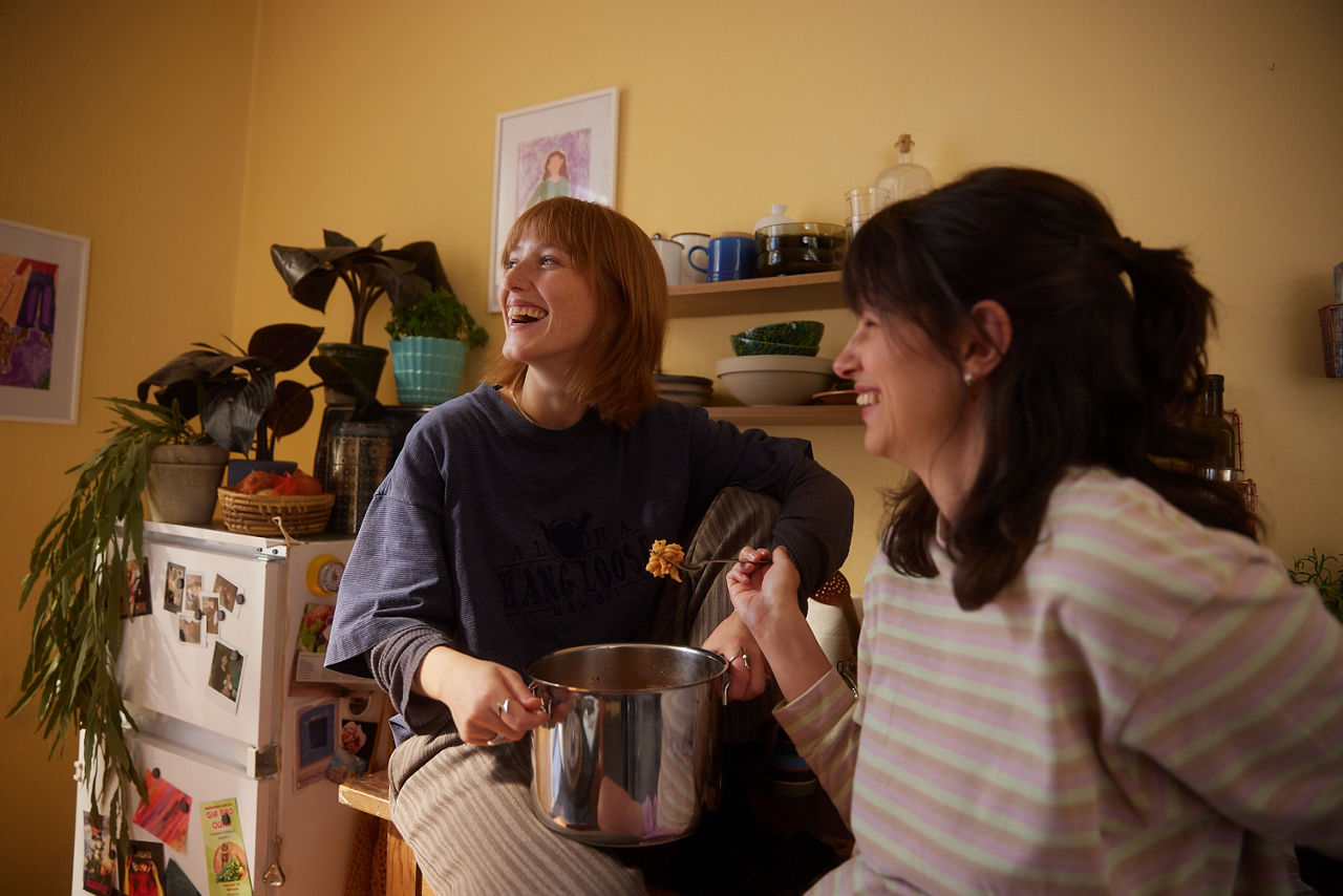 Zwei Freundinnen essen gemeinsam aus einem Topf Nudeln. Die Hintere sitzt auf der Küchenzeile, während sie die Frau im Vordergrund an die Küchenzeile anlehnt. 
